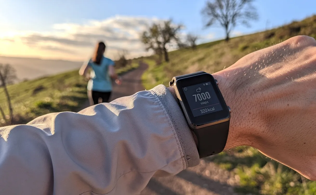 Bracelet connecté affichant 7000 pas et calories brûlées au poignet d’une personne marchant sur un sentier extérieur au coucher du soleil.