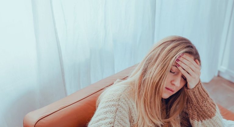 Jeune femme fatiguée et stressée assise sur un canapé, la main sur le front et les yeux fermés